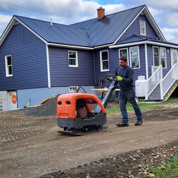 A man operates a compacting machine near a blue house under construction.