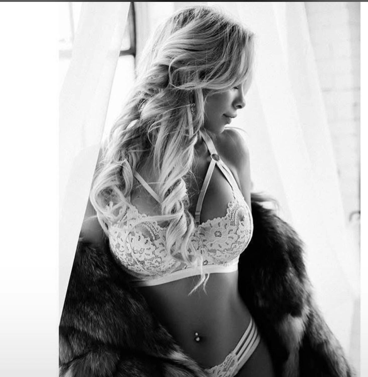 A woman with long wavy hair in lace lingerie and a fur coat poses by a window.