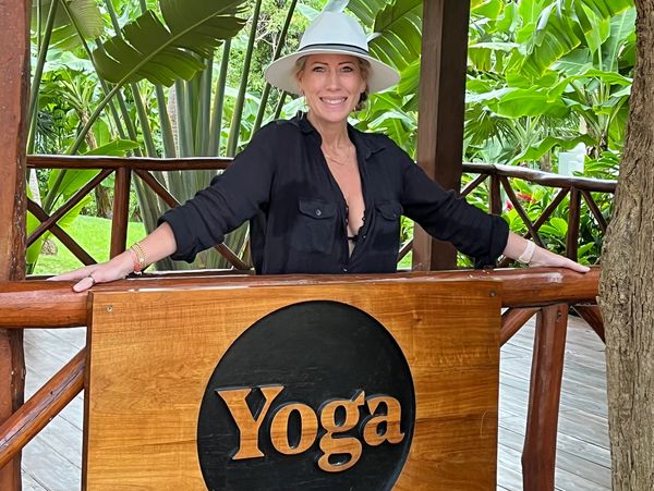 Woman smiling by a wooden yoga sign surrounded by tropical greenery.