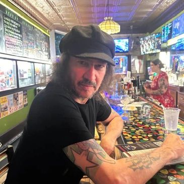 Man with tattoos sitting at a colorful bar counter wearing a black cap.