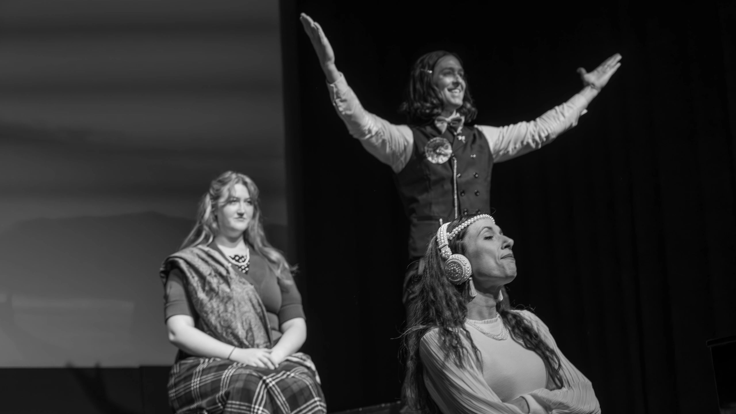 Three actors perform on stage in a dramatic black and white scene.