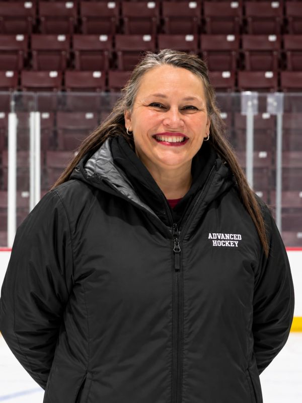 Smiling woman in a black 'Advanced Hockey' jacket at an ice rink.