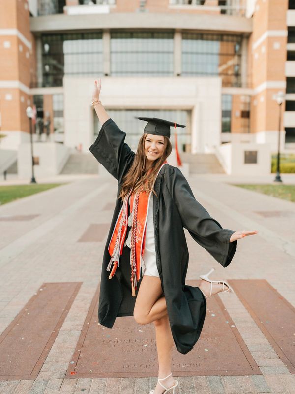 Happy female graduate posing in cap and gown outdoors with arms raised.