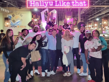 A smiling group of people posing in front of the Hallyu neon lights that read, "Hallyu Like That."