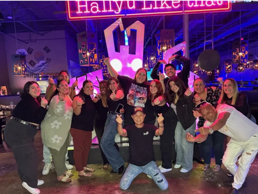 A group of smiling people standing in front of neon lights that read, "Hallyu Like That."