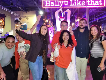 A group of smiling people standing in front of neon lights that read, "Hallyu Like That."