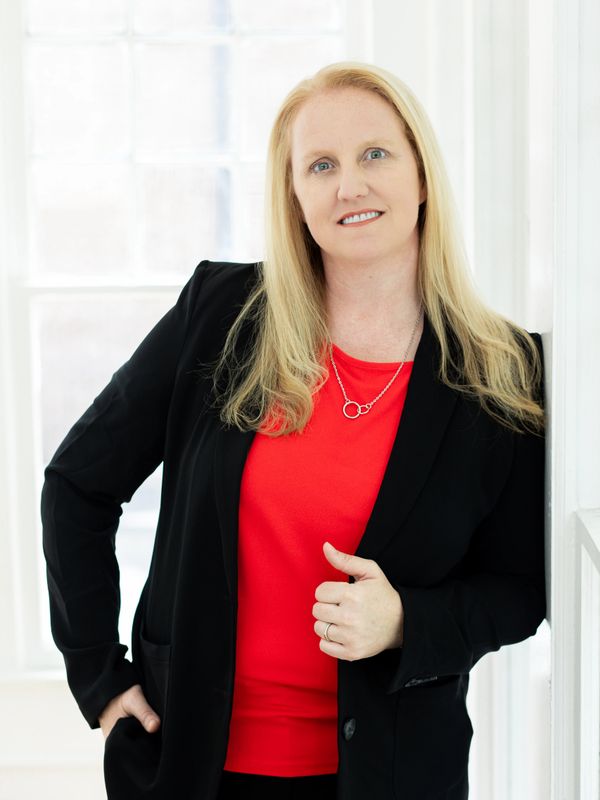 Professional woman in a black blazer and red top smiling by a window.