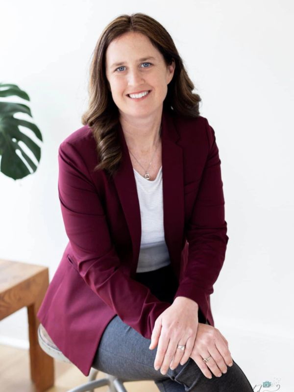 Smiling woman in a maroon blazer sitting on a stool indoors.