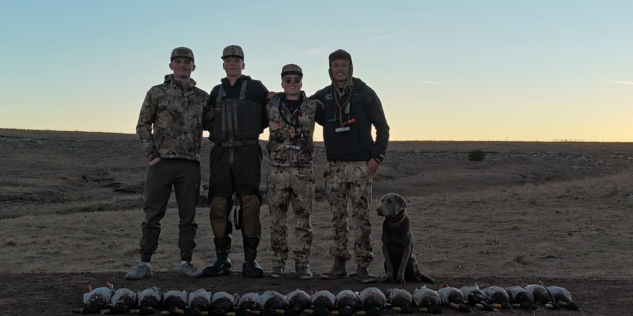 Four hunters in camouflage gear pose with their dog and hunted ducks at sunset.
