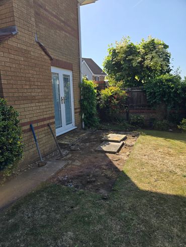 Backyard garden area beside a house with a patch of bare soil and garden tools.