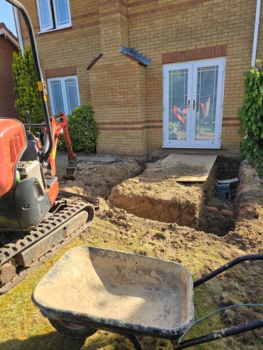 Excavation work in progress near a house entrance with a wheelbarrow and mini excavator.