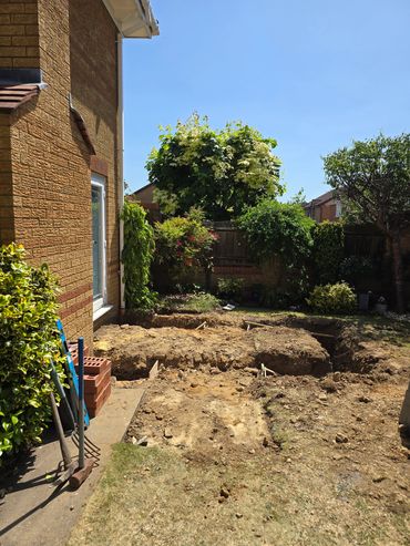 Backyard under construction with dug-up soil and gardening tools leaning against the house.