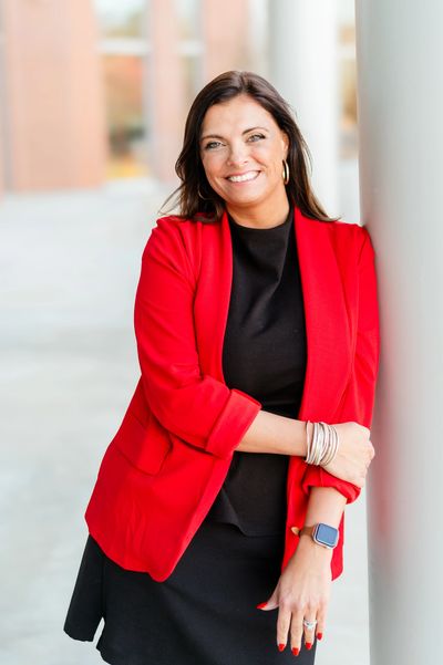 Confident woman in a red blazer smiling while leaning on a white pillar.