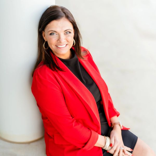 Smiling woman in red blazer sitting against a white pillar.