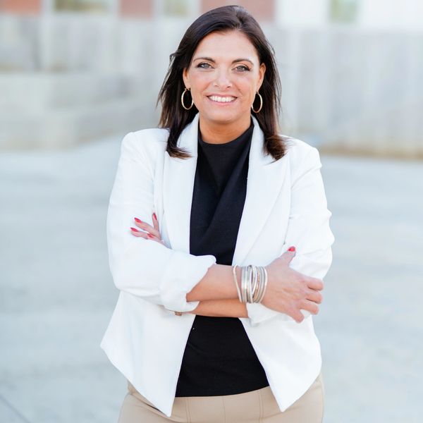 Confident woman in a white blazer and black top smiling with arms crossed outdoors.
