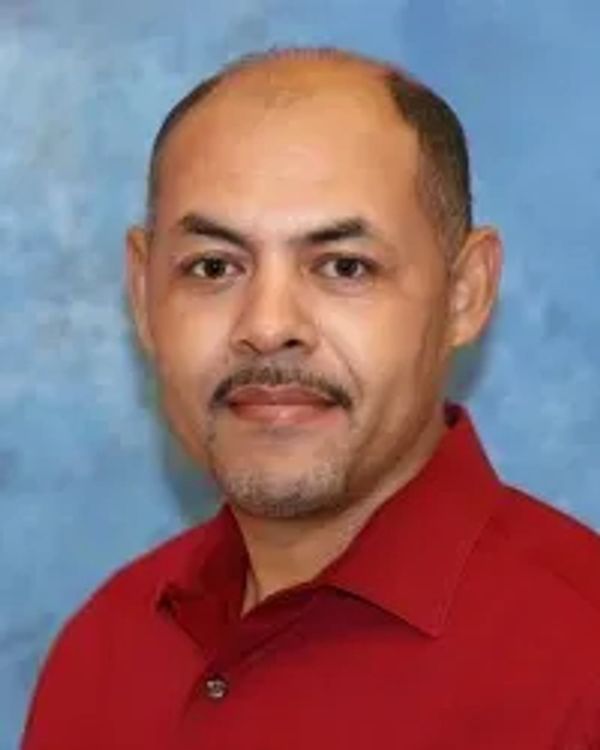 Man with short hair and mustache wearing a red collared shirt against a blue background.