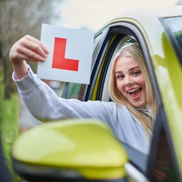 Happy young woman holding a learner driver L plate from a car window.
