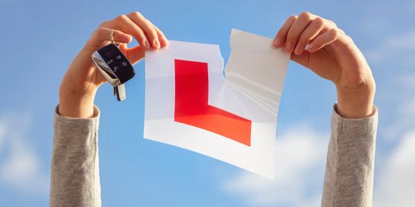 Hands holding car keys and a torn learner driver sign against blue sky.