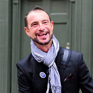 Smiling man with a scarf and blazer standing outdoors.