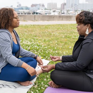 Two women sitting on mats outdoors, holding hands and facing each other.
