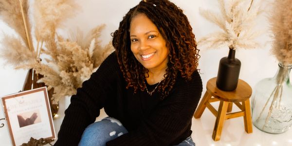 A smiling woman sits barefoot on the floor surrounded by decorative pampas grass and soft lights.