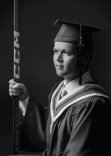 Graduate in cap and gown holding a hockey stick, looking thoughtful.