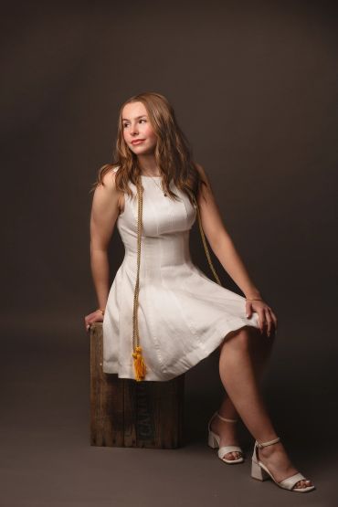 Young woman in a white dress sitting on a wooden box against a dark background.