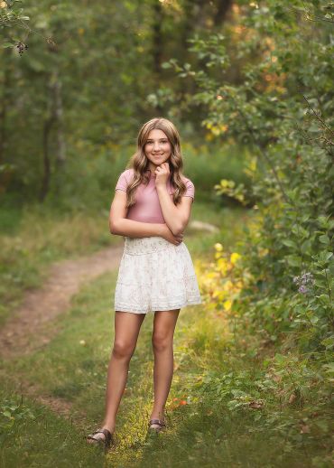 A young woman stands on a forest path, smiling and posing confidently.