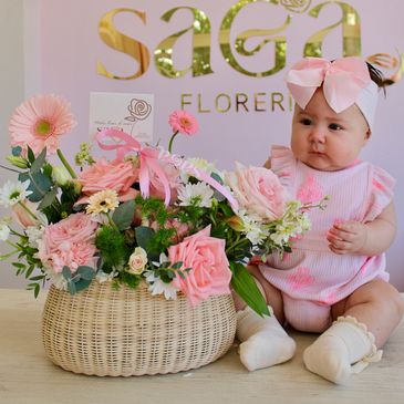 Bebé con diadema rosa junto a arreglo floral.