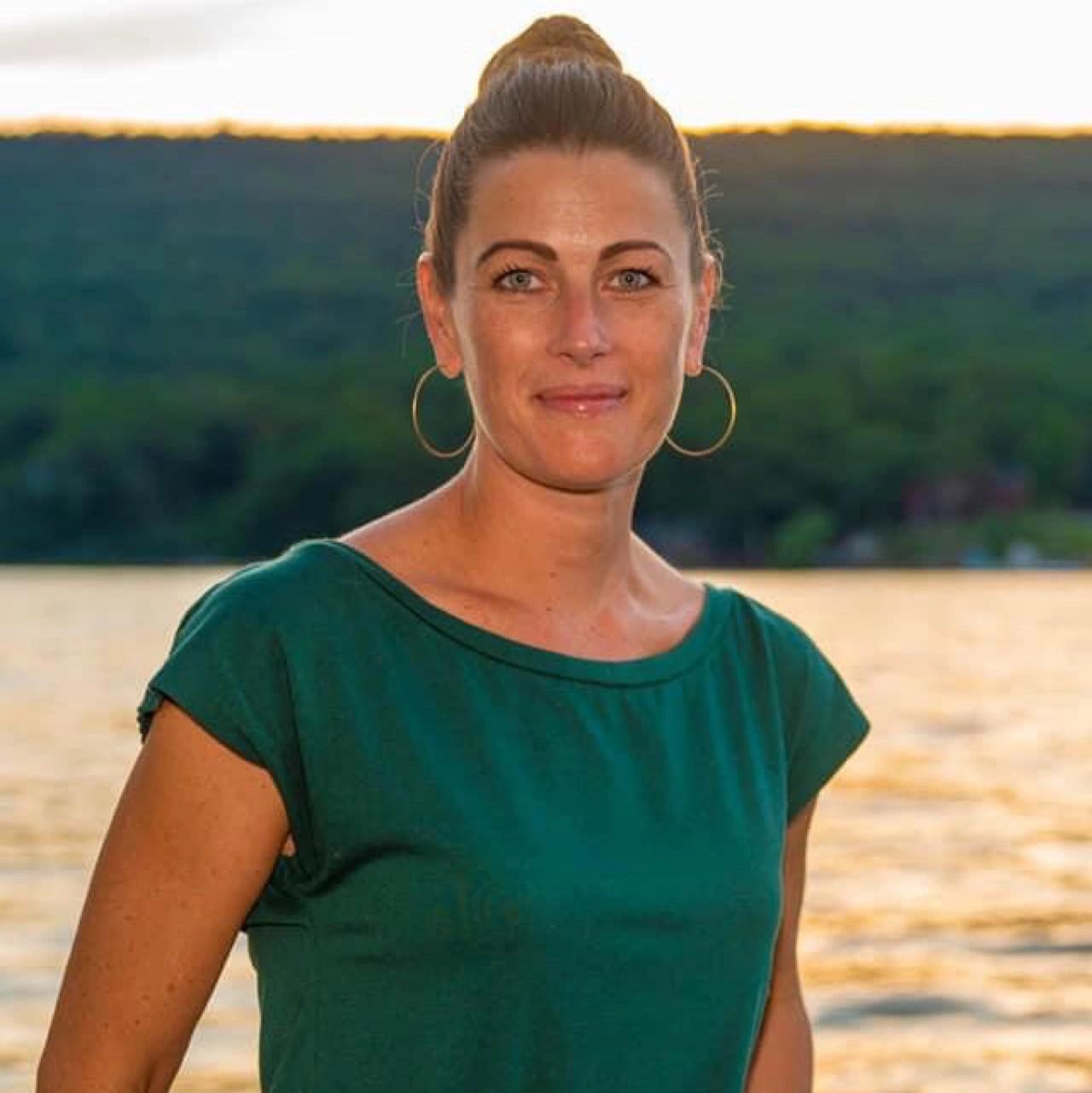 Woman in green top standing by a lake at sunset.