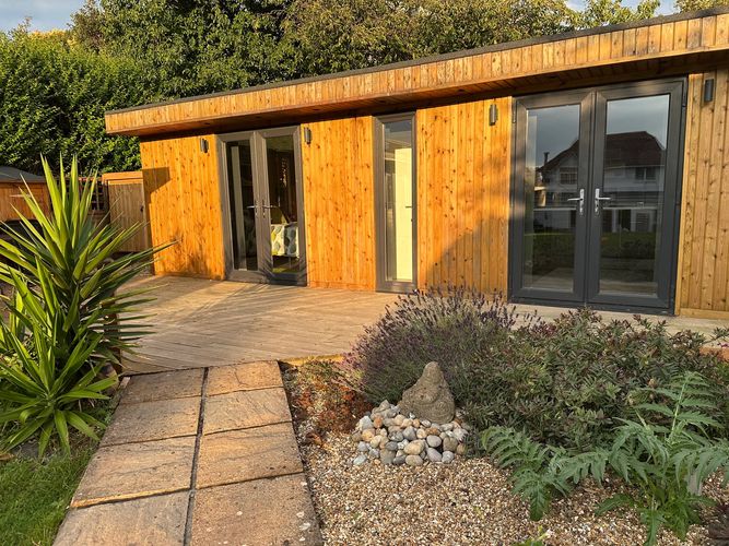 A wooden garden shed with glass doors and lush plants in front.