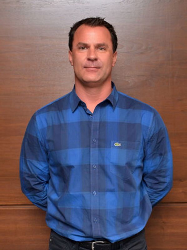 Man in a blue checkered shirt standing against a wooden wall.