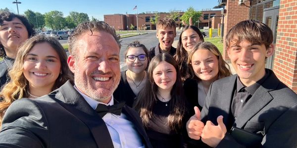 A group of smiling young people and a man in a tuxedo outdoors.