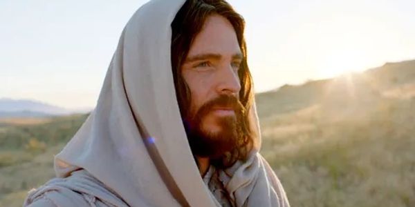 A man with a beard and long hair wearing a hooded robe in a sunlit landscape.