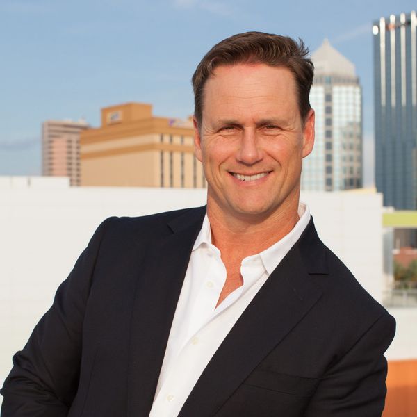 Man in a dark blazer and white shirt smiling outdoors in a city setting.