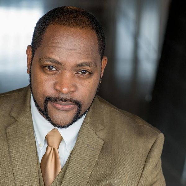 Confident man in a tan suit and gold tie looking at the camera.