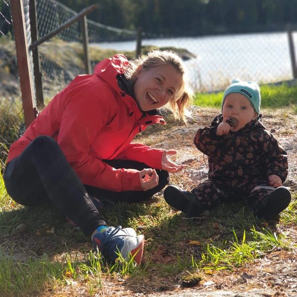 A picture of Kelsey in a red jacket, sitting on the grass with a young toddler eating a pinecone.