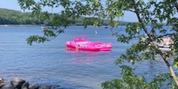 A bright pink inflatable float on a calm lake near a rocky shore and leafy tree.