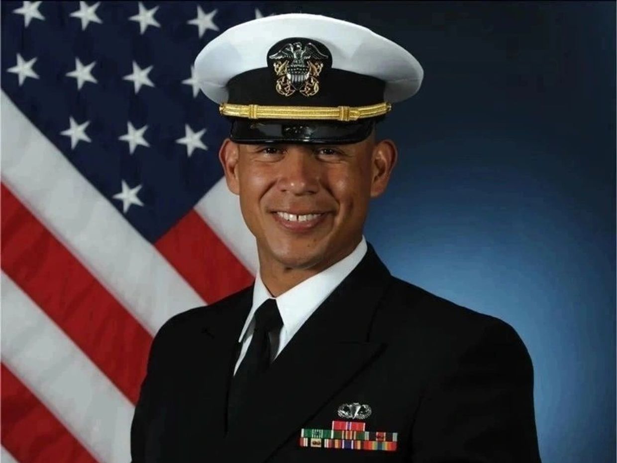 Smiling U.S. Navy officer in uniform with flag background.