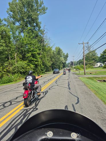 Motorcyclists riding on a tree-lined suburban road on a sunny day.