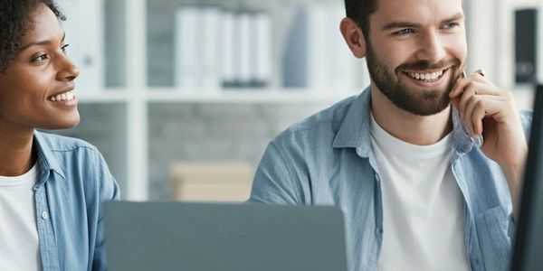 Two colleagues smiling and working together at a desk with laptops.