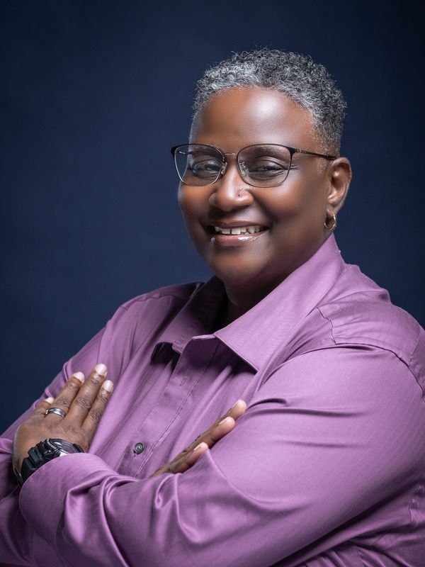 Confident person with short gray hair and glasses smiling warmly in a purple shirt.