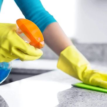 Person cleaning countertop with spray bottle and cloth wearing yellow gloves.