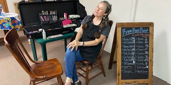 Woman sitting next to face painting supplies and a menu board for free face painting.