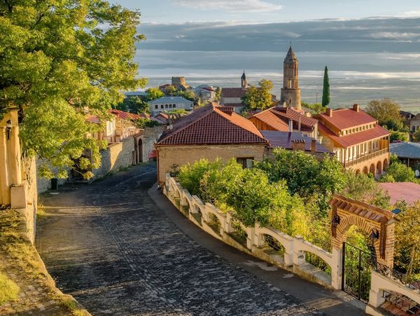 A quaint village street with red-roofed buildings and lush greenery under a partly cloudy sky.