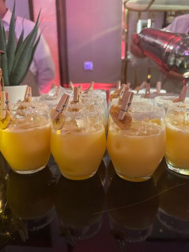 Glasses of orange cocktails with ice and decorative clips on a bar counter.