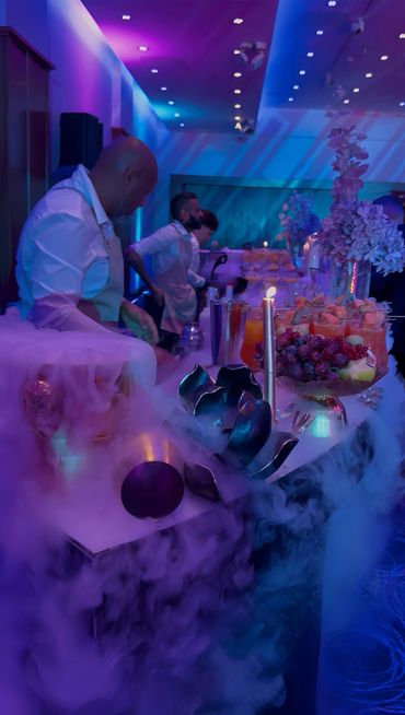Bartenders serve drinks at a vibrant, smoke-filled event with colorful lighting.
