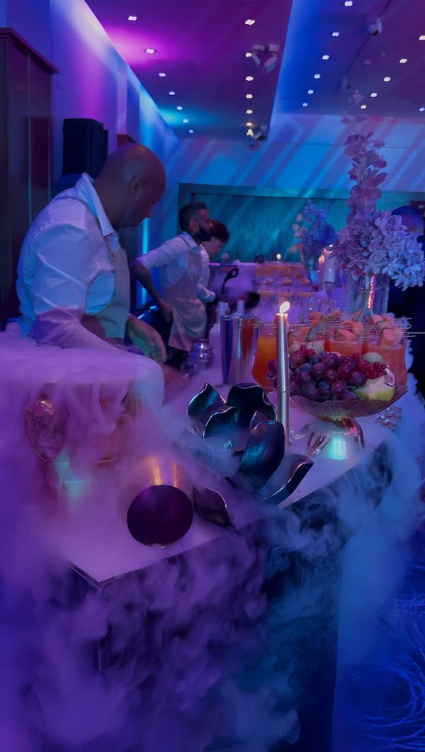 Bartenders serve drinks at a vibrant, smoke-filled event with colorful lighting.