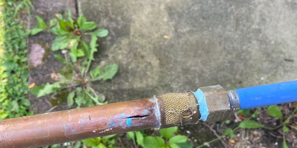 Cracked copper pipe with a blue hose attached outdoors.