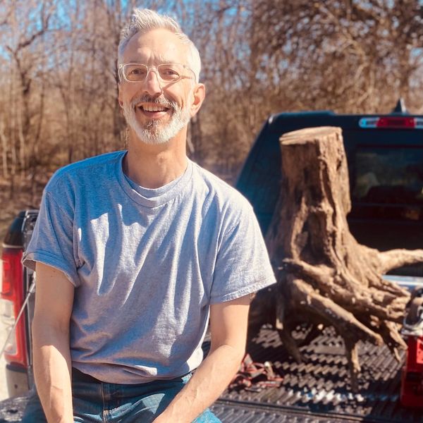 Smiling man with glasses sitting on a truck bed with a large tree stump behind him.